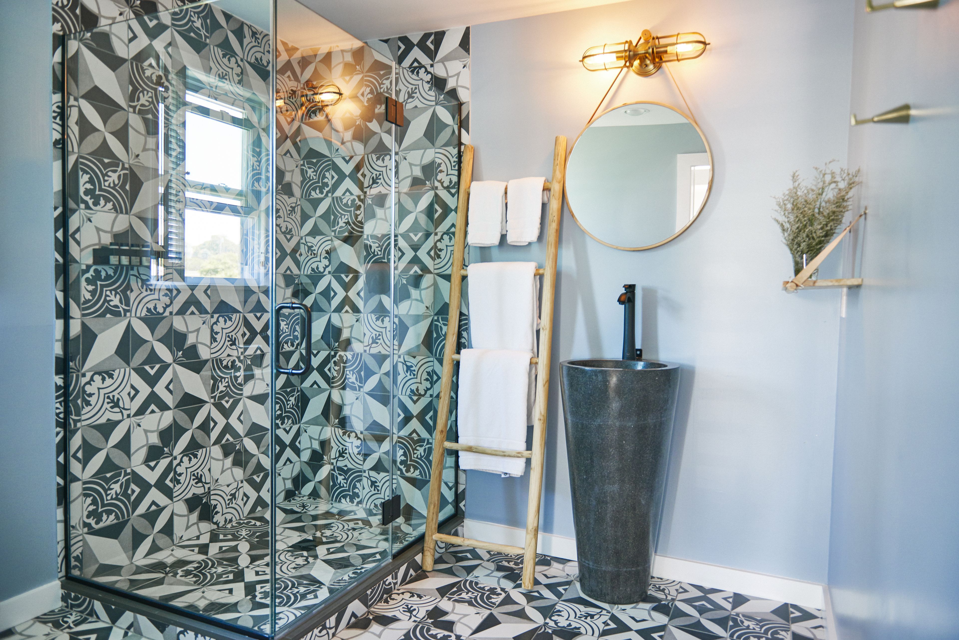 A bathroom with patterned tiles, a glass shower enclosure, a wooden ladder, a round mirror, and a black sink.