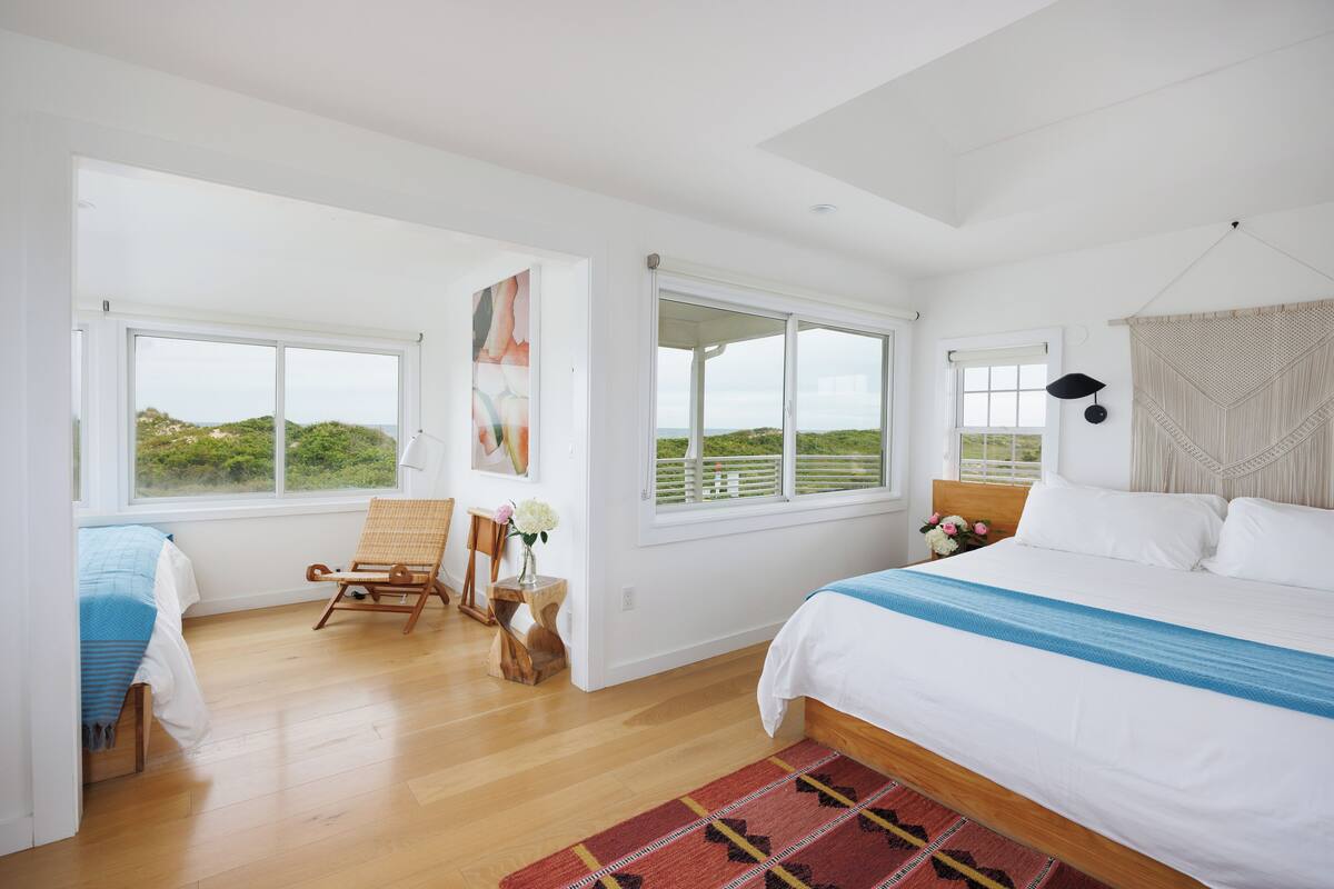 A bedroom with a large bed, a chair, a small table with flowers, and a view of the outdoors through large windows.
