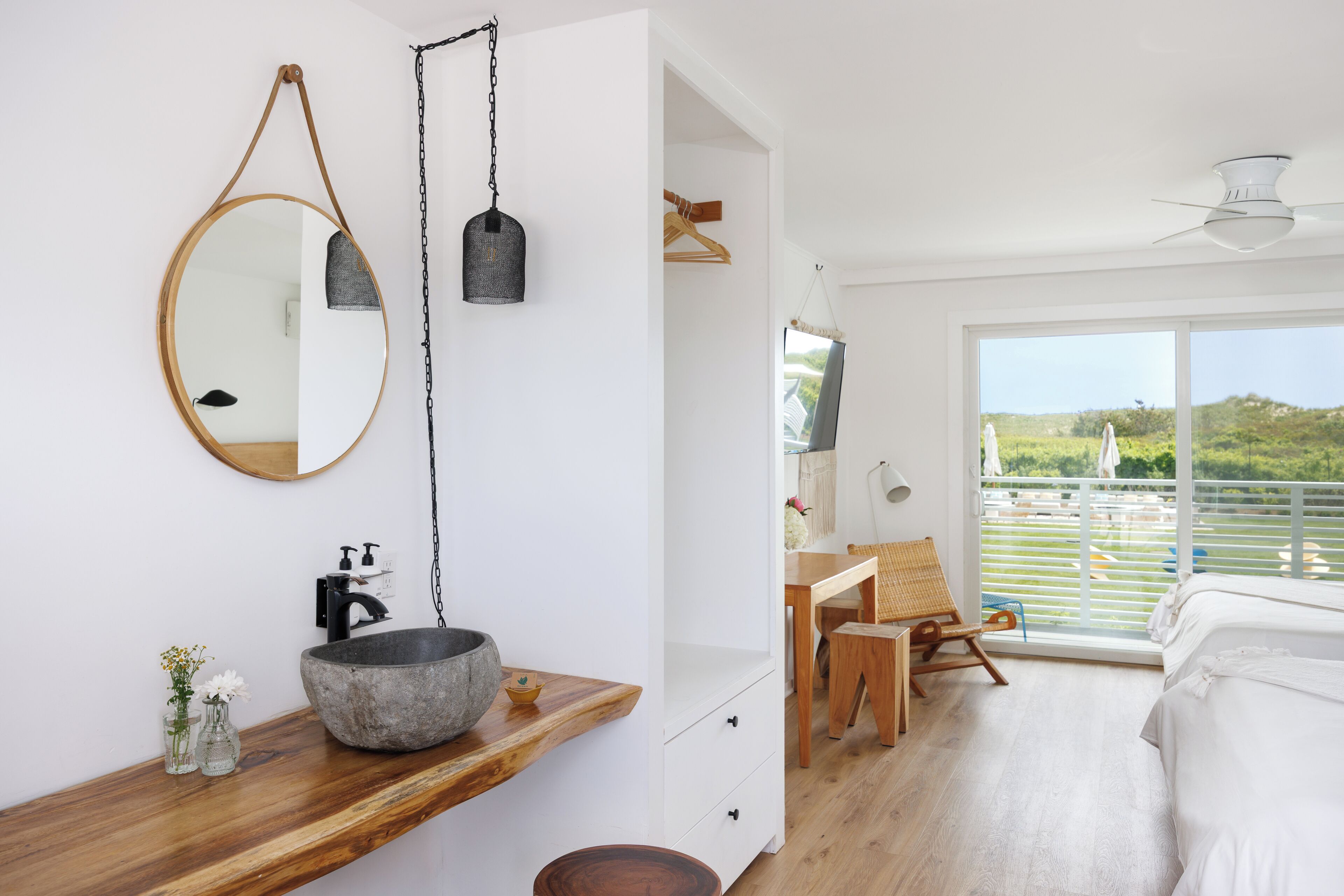A modern bedroom with a large window, a wooden bedside table, a round mirror, and a hanging light fixture.