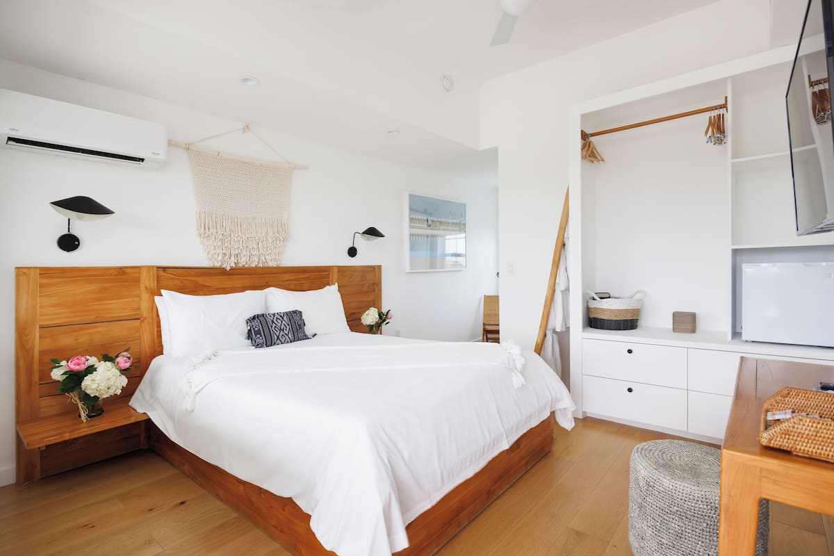 A modern bedroom with a wooden bed, white bedding, a small bedside table with flowers, a wall-mounted air conditioner, and a TV on a stand.