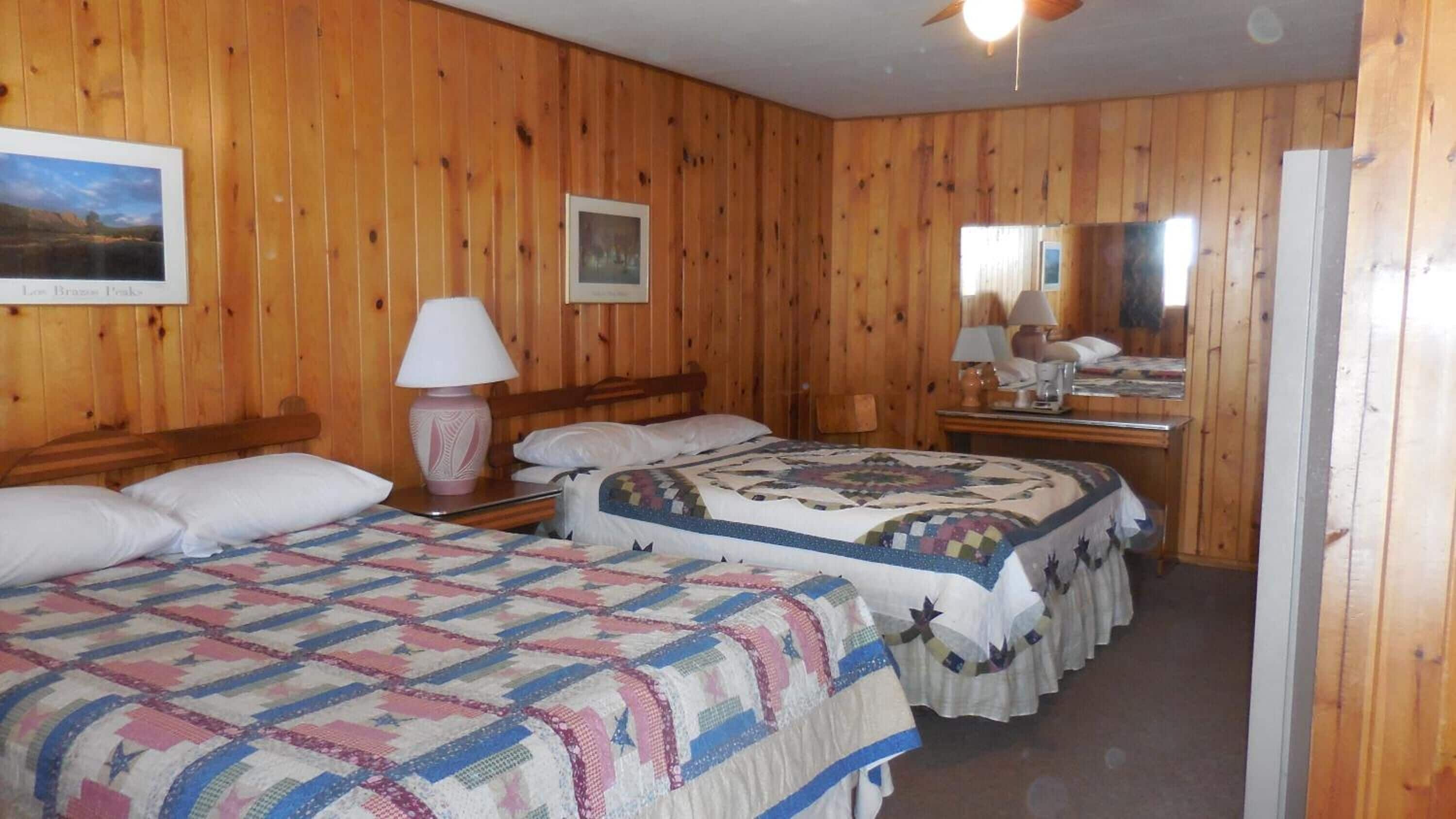 Una habitación con paneles de madera, con dos camas, cada una con colcha, una mesita de noche con lámpara y un espejo en la pared.
