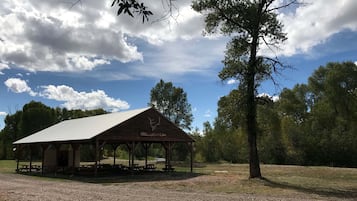 Un pabellón de madera rústica con techo inclinado, identificado como "Elk High Lodge", rodeado de árboles y un cielo despejado.