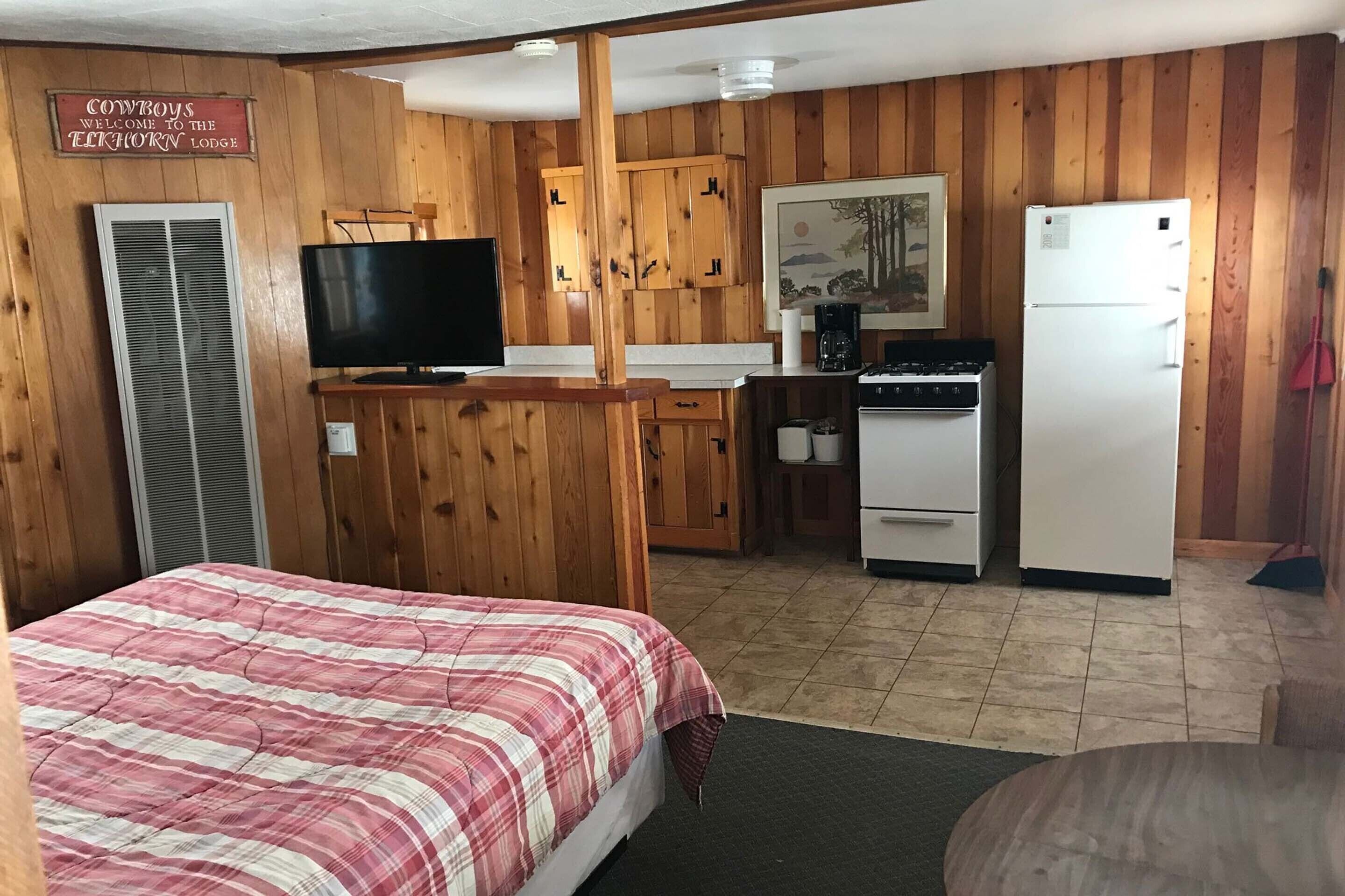A room with a bed, a television, wooden cabinets, a refrigerator, and a kitchen area.