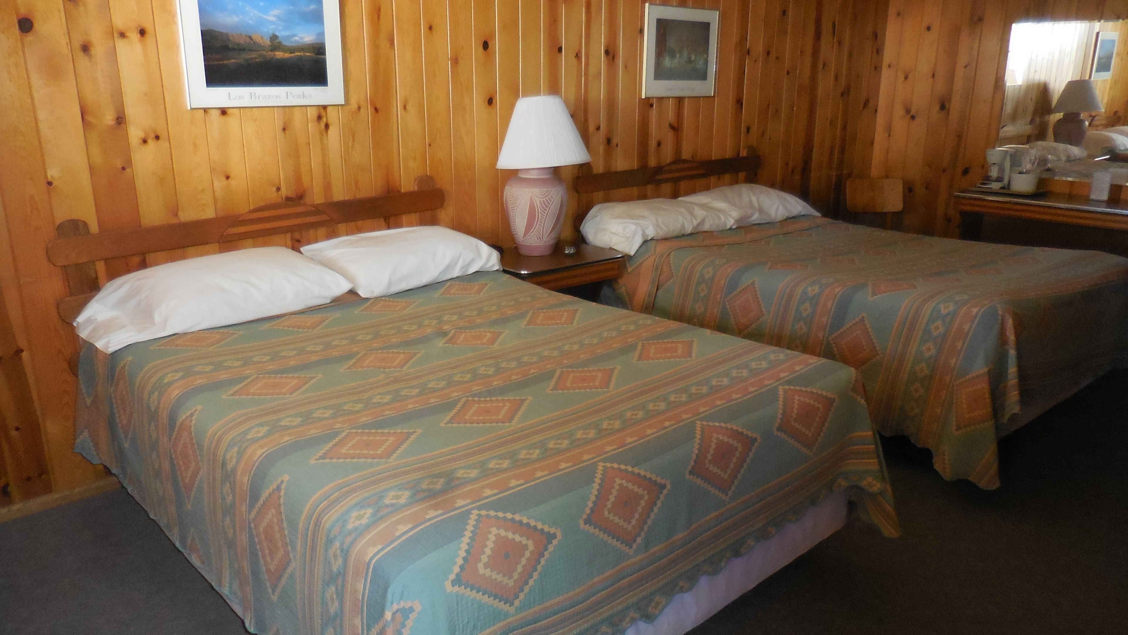 A room with two beds, wooden walls, a nightstand with a lamp, and framed pictures on the wall.