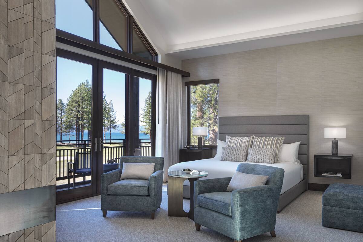 A modern hotel room with a large bed, two armchairs, a small table, and a view of the outdoors.