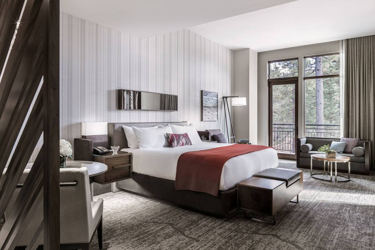 A modern hotel room with a large bed, a sofa, a small table, and a nightstand. There is a window with a view of trees and a balcony.