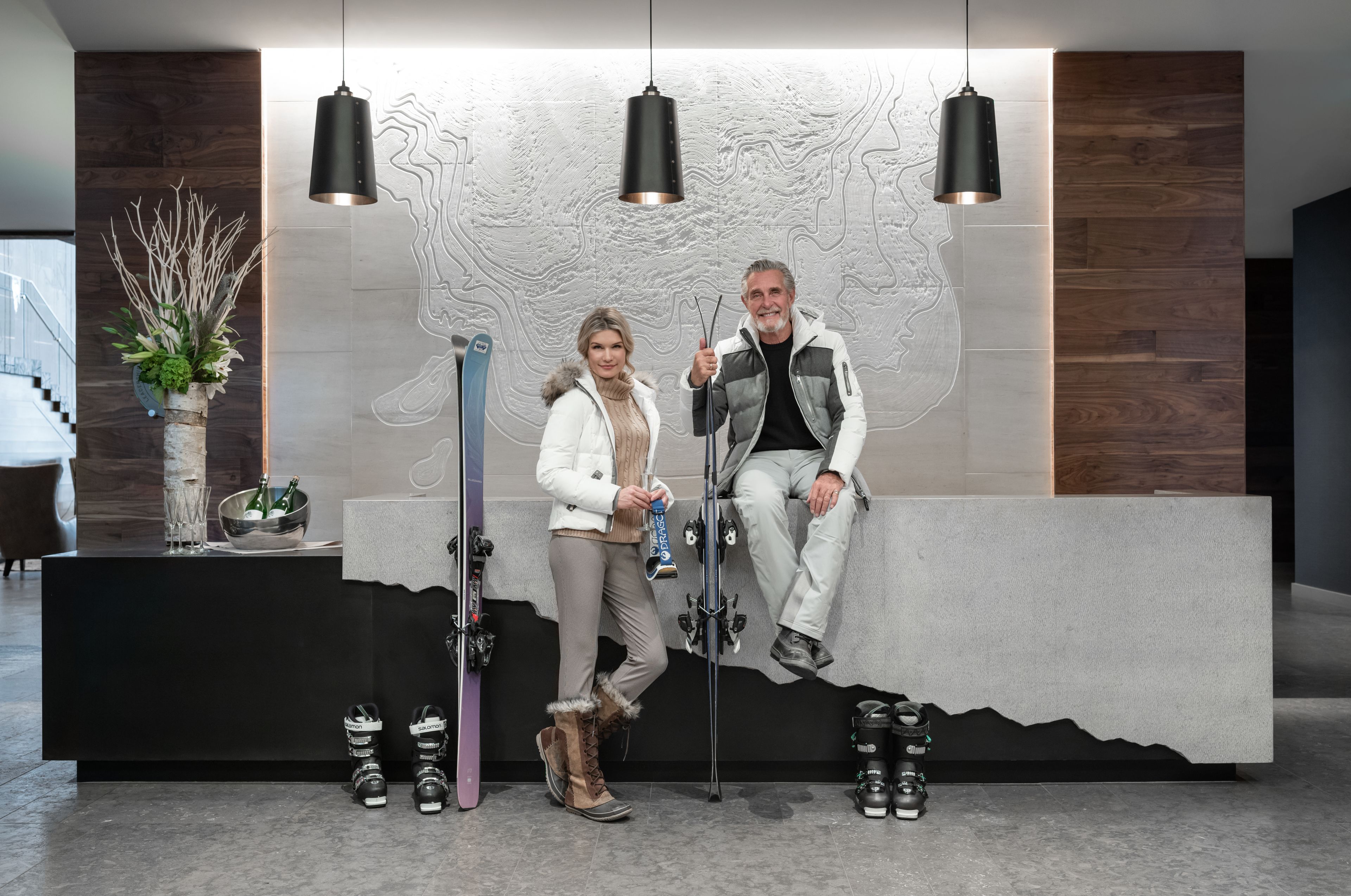 Een modern ontworpen receptiegebied met een receptiebalie, twee personen zittend, ski's en een sierplant.