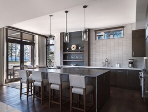 A modern kitchen with a large island, bar stools, pendant lighting, and a view through the glass door.
