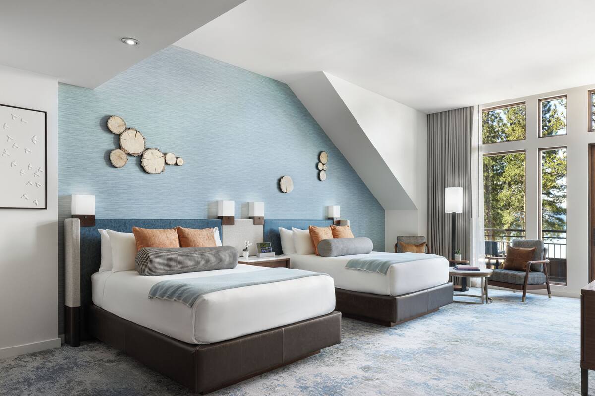 A modern hotel room with two beds, a large window, and a view of trees.