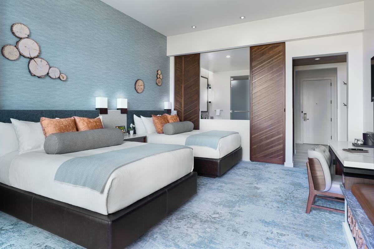A modern hotel room with a large bed, two smaller beds, a desk, and a bathroom visible through an open door.