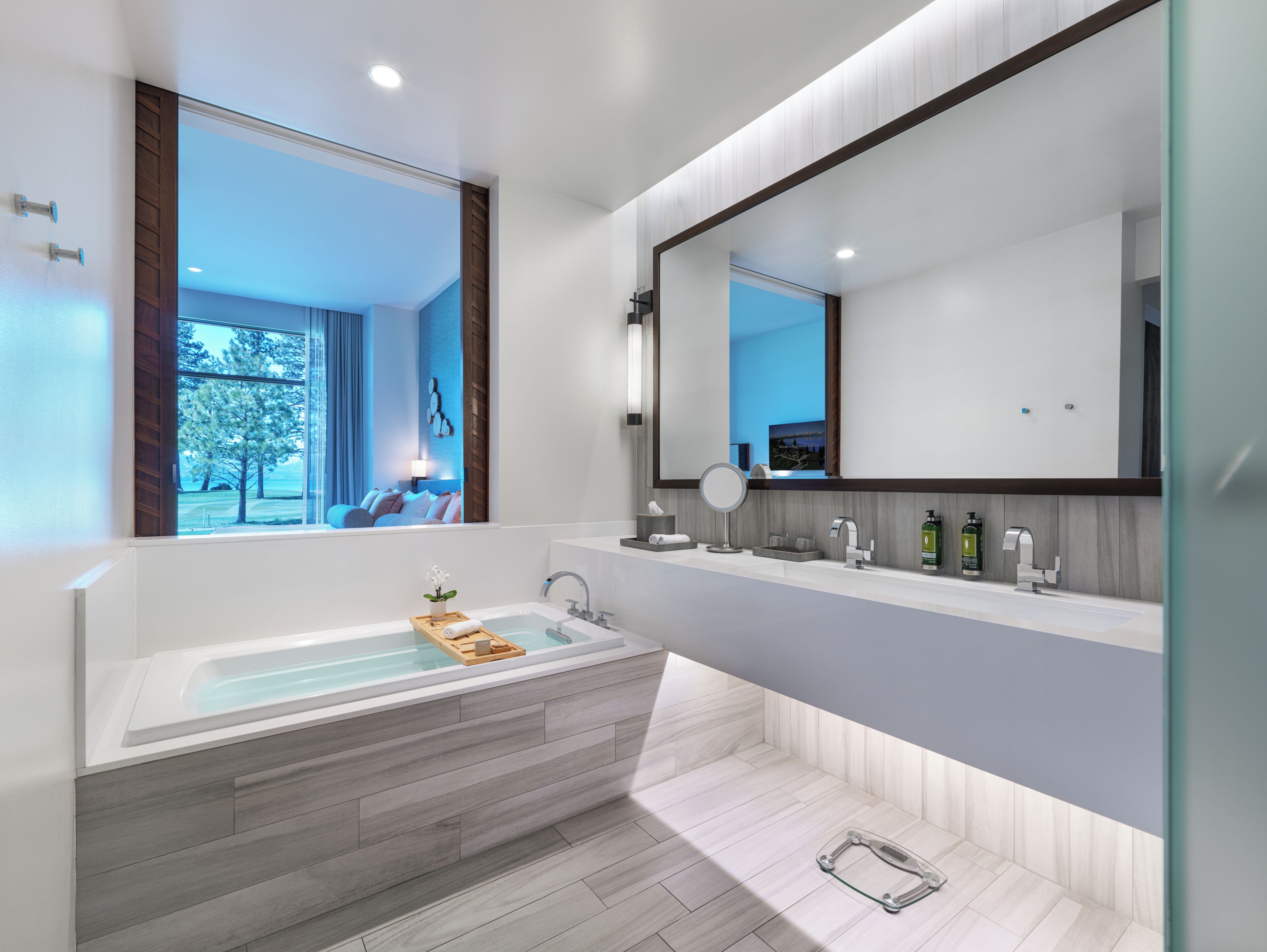 A modern bathroom with a large bathtub, a vanity with a mirror, and a view of a bedroom through a window.