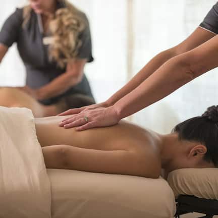 A massage therapy session in a room with a massage table, a person receiving a back massage, and another person assisting.