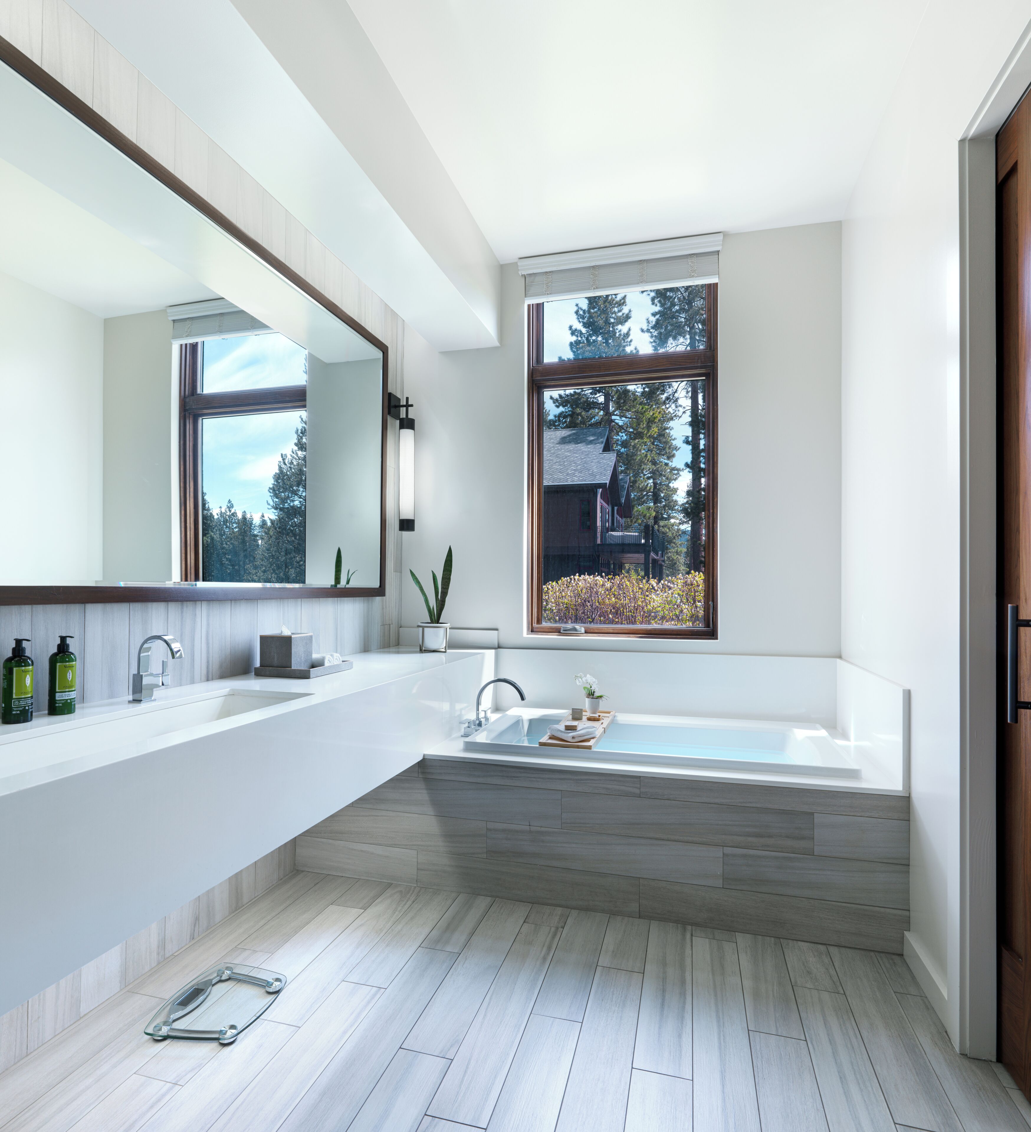 Un baño moderno con un espejo grande, dos lavamanos, una bañera y una ventana con vistas a árboles.