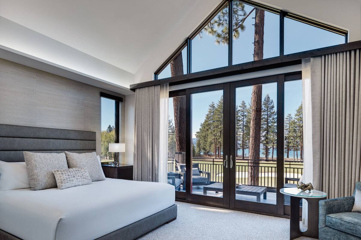 A modern hotel room with a large bed, a seating area with a view of the outdoors, and a balcony with a glass door.
