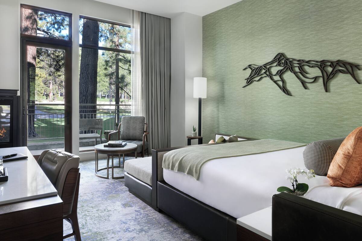 A hotel room with a bed, a desk, a chair, a lamp, and a large window with a view of trees.