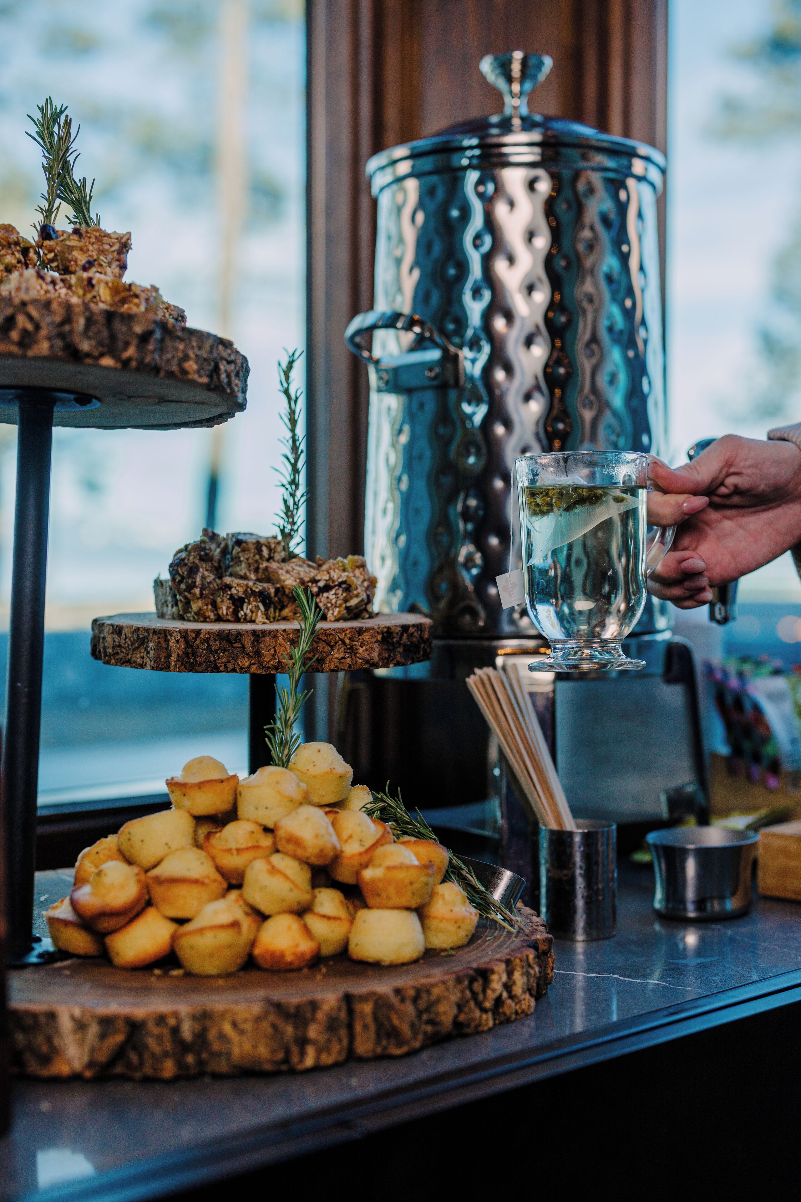 A buffet setup with roasted potatoes, a dish with rosemary, and a large metal container in the background.