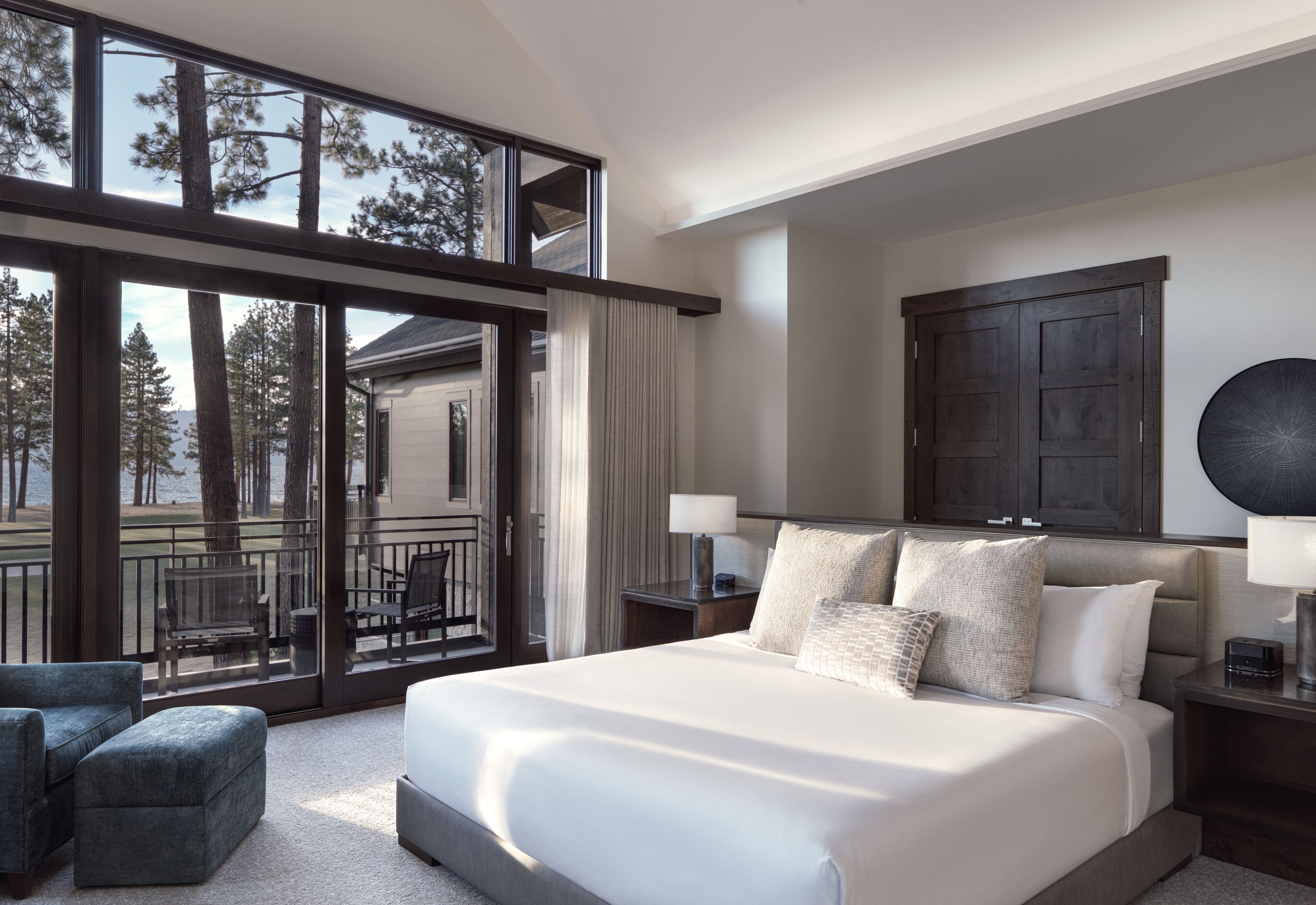 A modern bedroom with a large bed, two bedside tables, a view of trees through floor-to-ceiling windows, and a balcony with outdoor furniture.