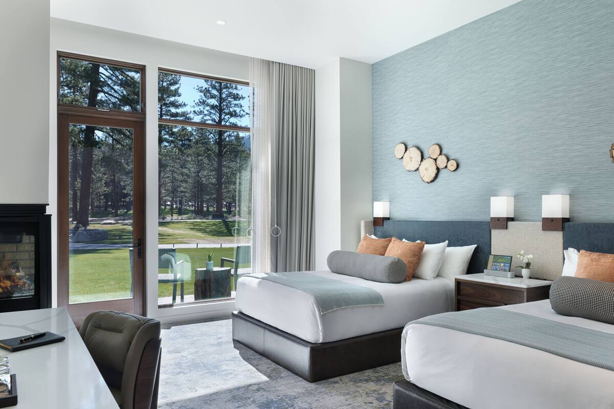 A hotel room with a large sliding glass door, a bed with a gray and white color scheme, a nightstand with a lamp, and a wall-mounted light fixture.