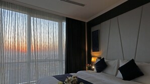 Een hotelkamer met een groot bed, een nachtkastje met een lamp en uitzicht op de ondergaande zon door dunne gordijnen.