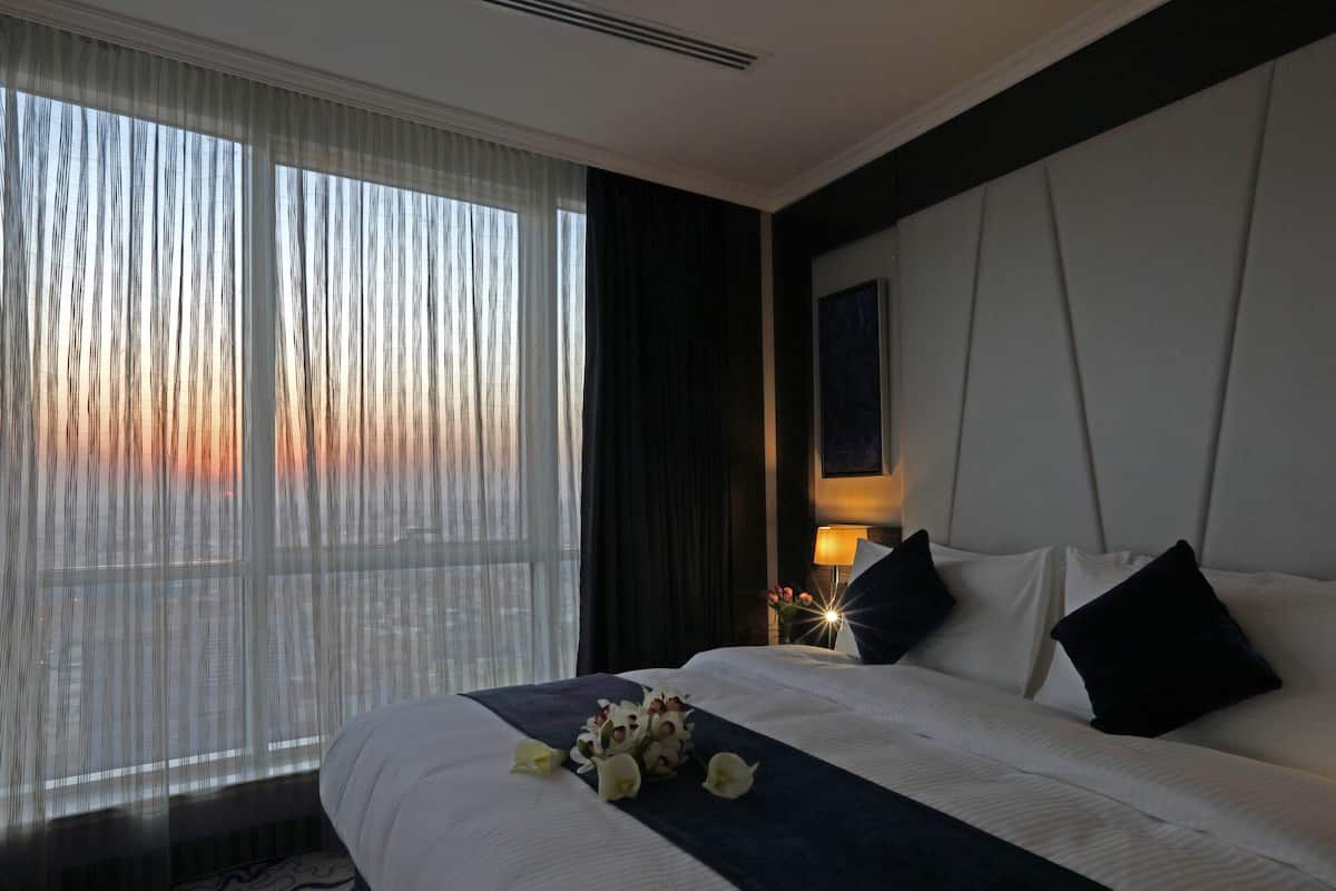 Hotellihuone, jossa on suuri sänky, yöpöytä lampun kanssa ja näkymä auringonlaskuun ohuiden verhojen läpi.