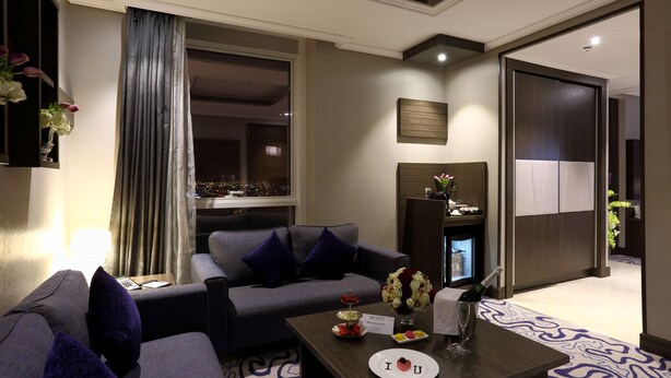 Een moderne hotelkamer met een grijze bank, een klein tafeltje met een bord, een bloevas en een minibar.