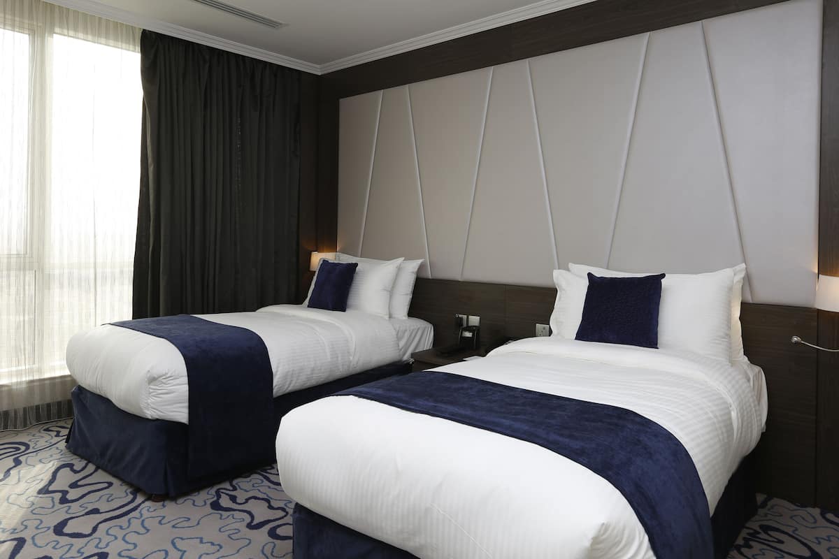 Hotellihuone, jossa on kaksi sänkyä, suuri ikkuna, jossa on verhot, kuviollinen matto ja moderni sängynpääty.
