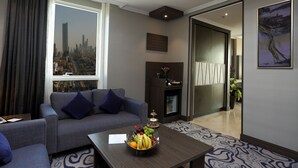 Een moderne hotelkamer met een grijze bank, een bijzettafeltje met fruit, uitzicht over de stad en een groot schilderij aan de muur.