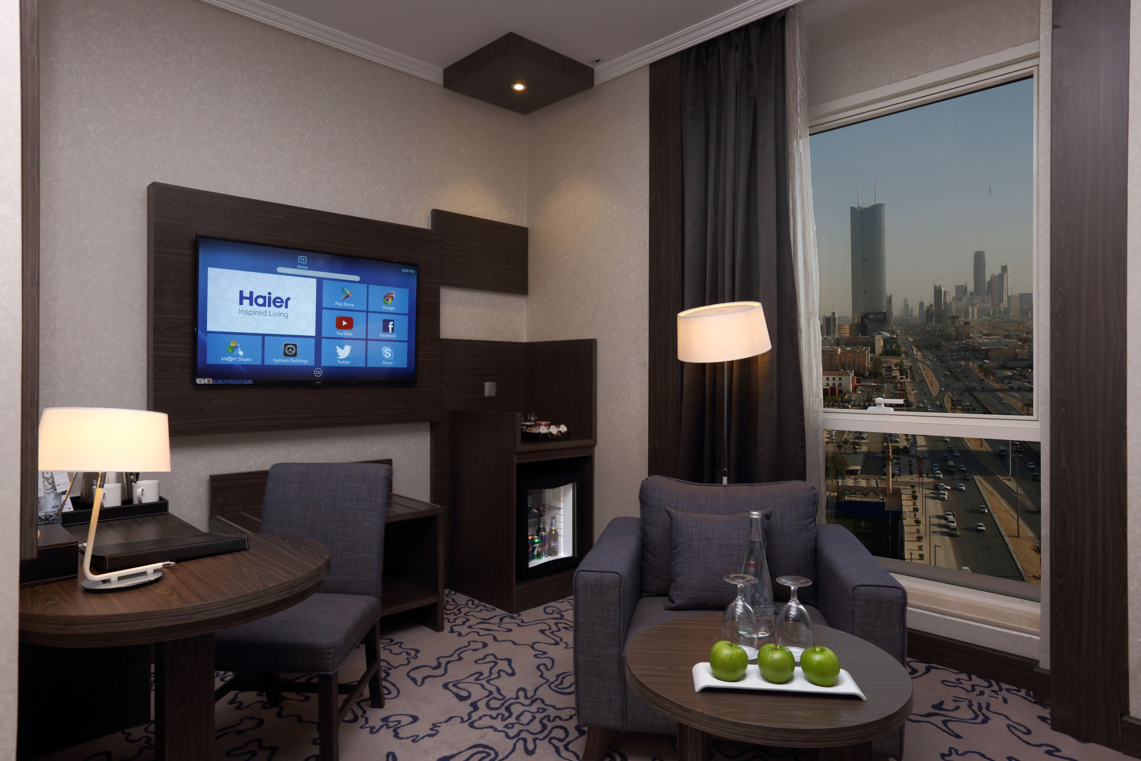A hotel room with a flat-screen TV mounted on the wall, a sofa, a small table with a tray of apples, and a large window offering a city view.