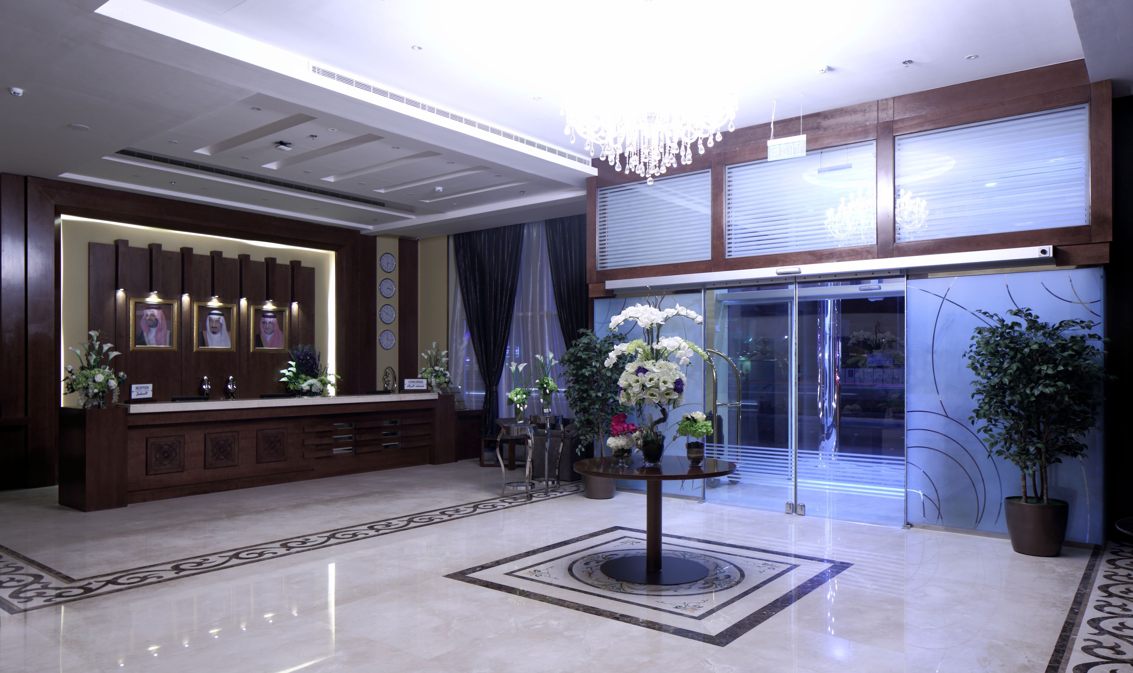 A hotel lobby with a reception desk, a chandelier, a clock, and a large window with blinds.