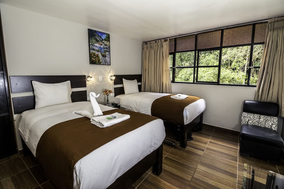 A hotel room with two beds, a large window with curtains, a painting on the wall, and a chair with a patterned cushion.