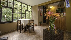 Ein Essbereich mit Tisch und Stühlen, ein Fenster mit Blick ins Grüne, eine Vase mit Blumen auf einem Tisch und eine Uhr an der Wand.