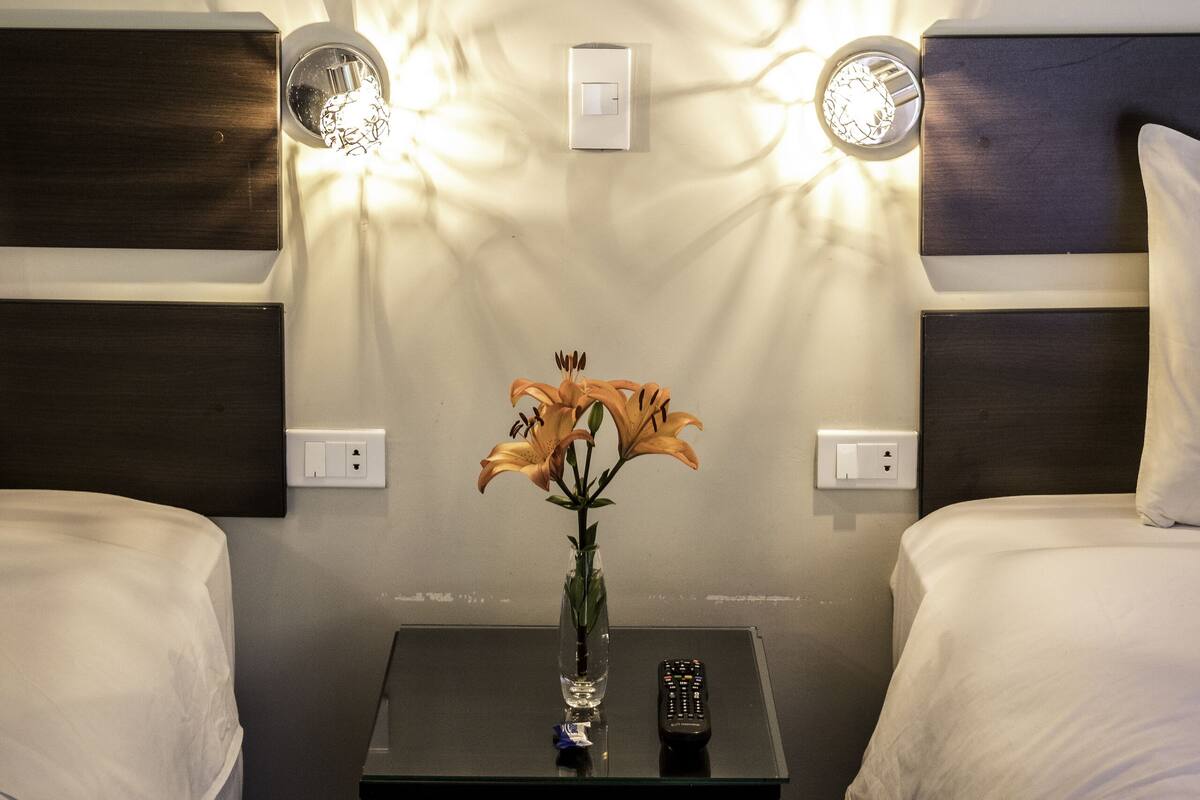A hotel room with two beds, a nightstand with a vase of flowers, a painting on the wall, and wall-mounted lights.