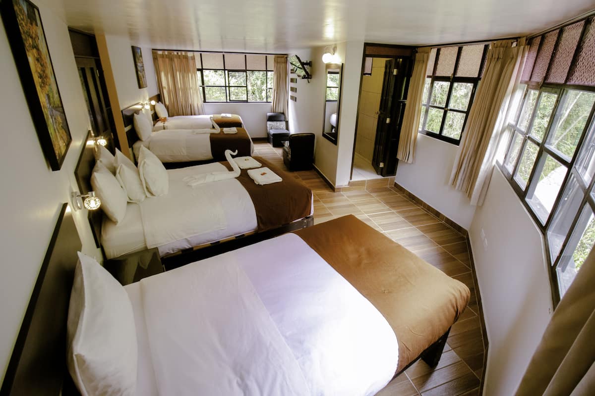 A hotel room with three beds, a small desk, and large windows.