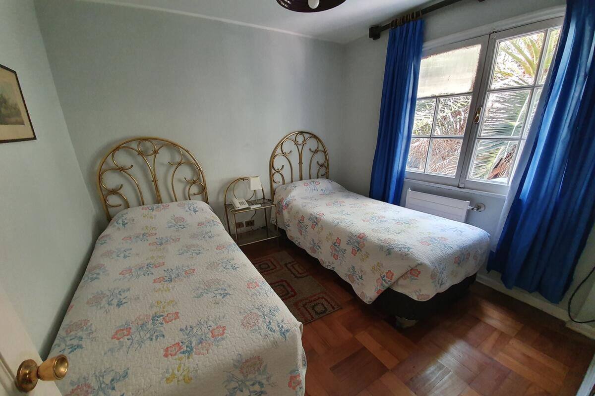 A bedroom with two single beds, a small bedside table with a lamp, a window with blue curtains, and a radiator.