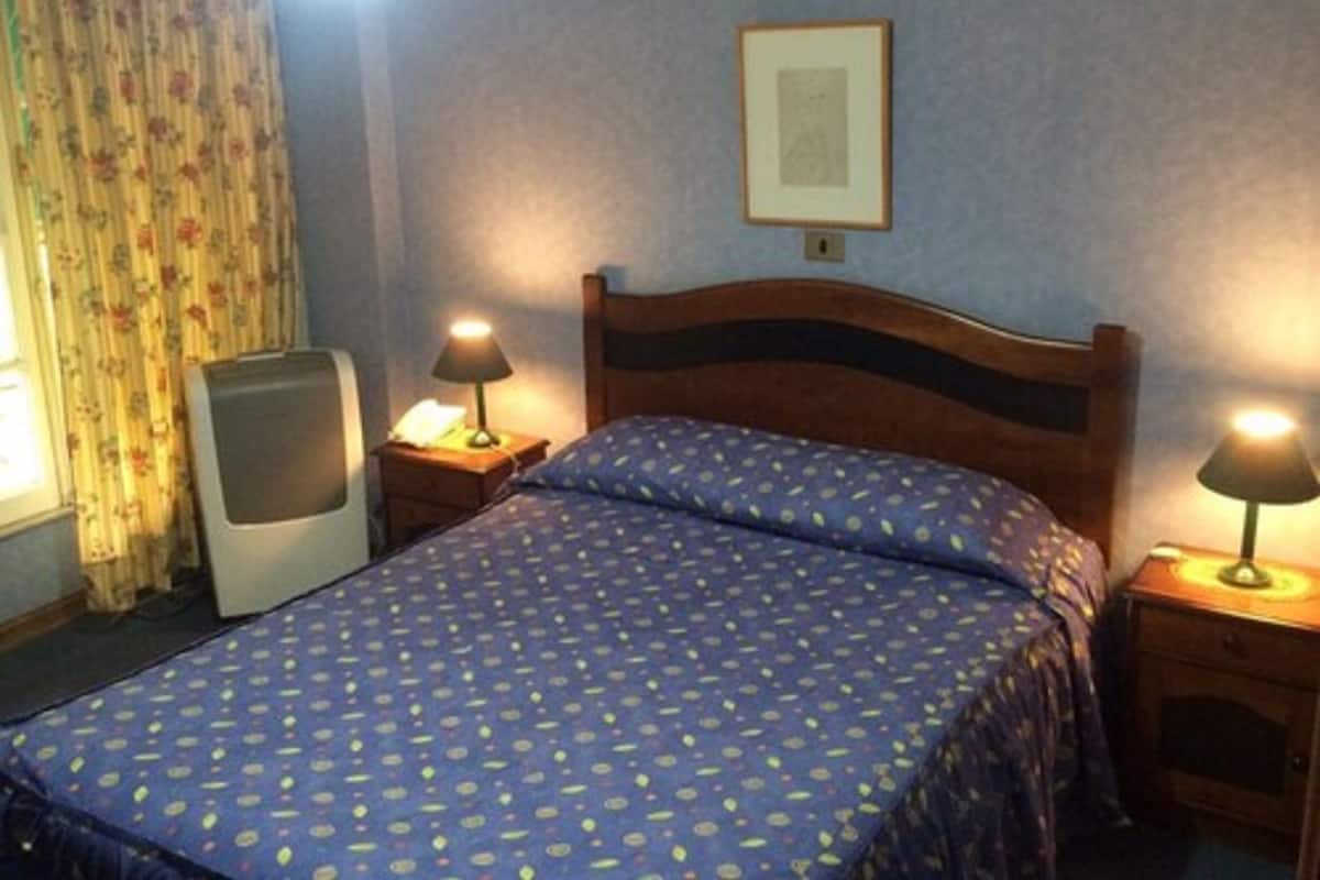 A bedroom with a bed, two bedside lamps, a framed picture on the wall, and a window with curtains.
