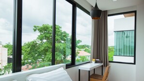 A modern room with a large window, a white bed, a wooden chair, and a small table with a plant.