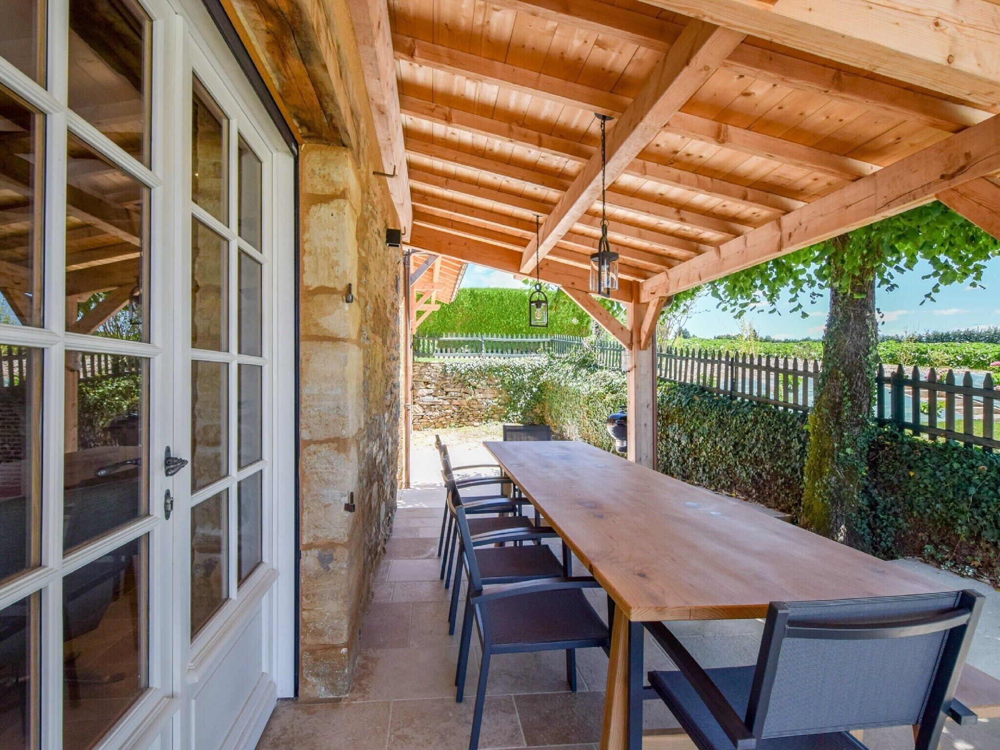 Une terrasse couverte avec une table et des chaises en bois, des murs en pierre et une vue sur un jardin et une clôture.