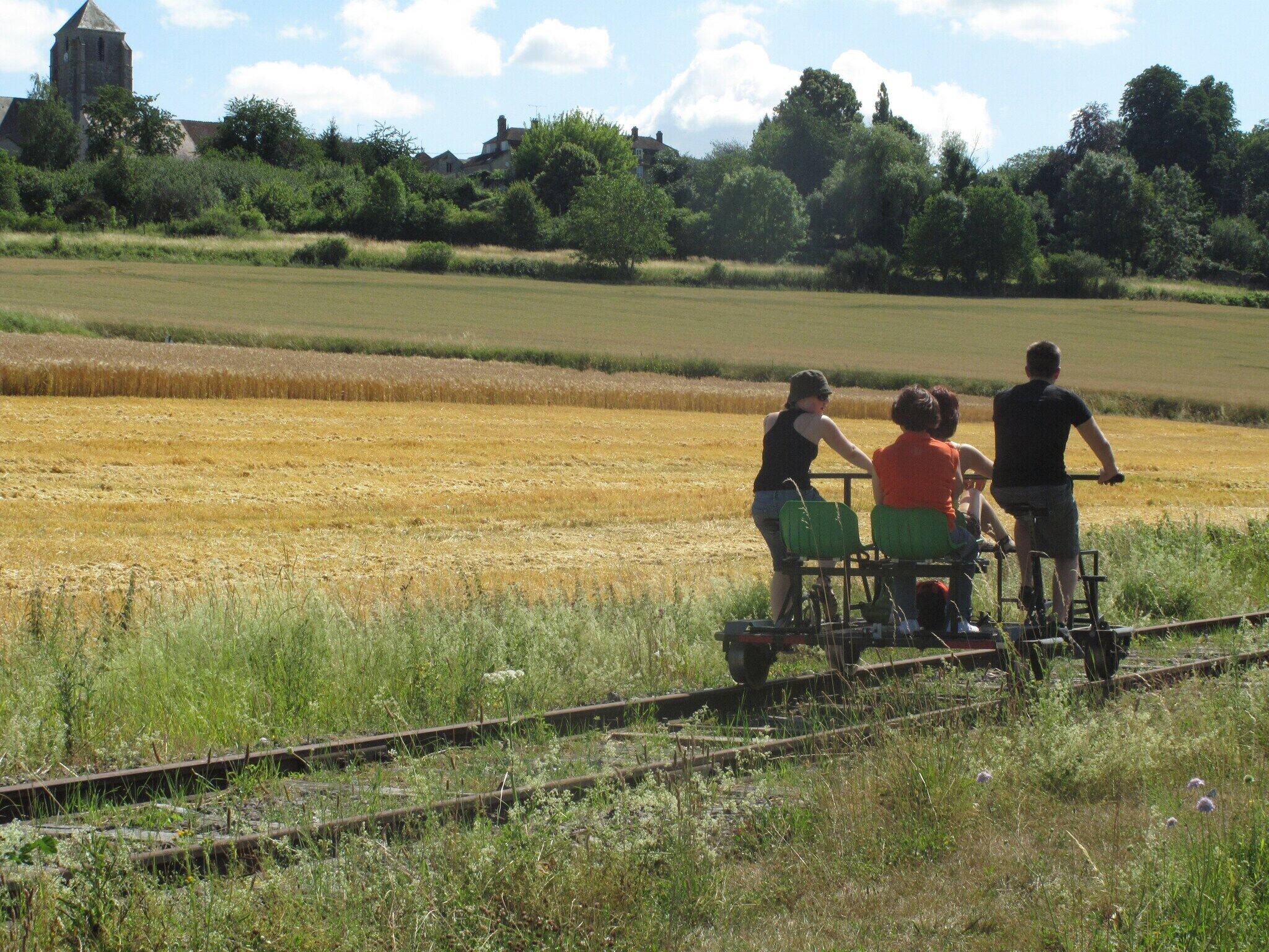 Eine Gruppe von Menschen in einem Zugabteil in ländlicher Umgebung, mit Feldern und einem Gebäude im Hintergrund.