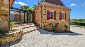 Une maison en pierre avec un toit en pente et des volets rouges, une allée gravillonnée et une véranda donnant sur un espace abrité.