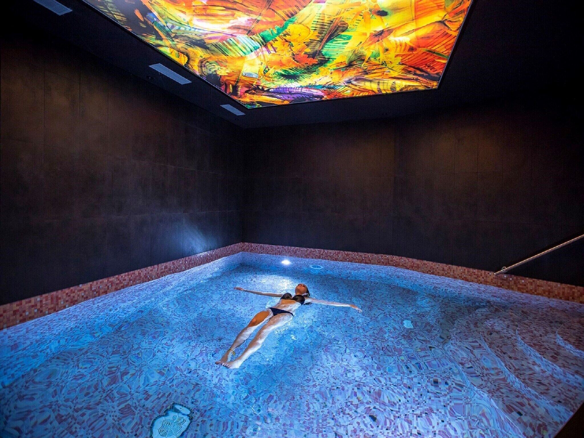 A person floating in a large indoor pool with a colorful ceiling.