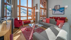 Ein modernes Wohnzimmer mit einer Couch, roten Sesseln, einem Glastisch und großen Fenstern mit Blick auf einen verschneiten Berg.