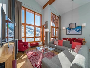Ein modernes Wohnzimmer mit einer Couch, roten Sesseln, einem Glastisch und großen Fenstern mit Blick auf einen verschneiten Berg.