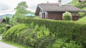 Et rustikt hus med tegltag, omgivet af frodig vegetation og en hæk.