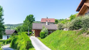 En landevej, der fører til et hus med røde tegltage, omgivet af frodig vegetation og bakker i baggrunden.