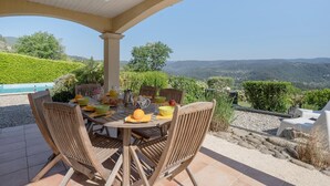 Eine überdachte Terrasse mit Esstisch und Stühlen, mit Blick auf eine malerische Landschaft aus Hügeln und Bäumen.