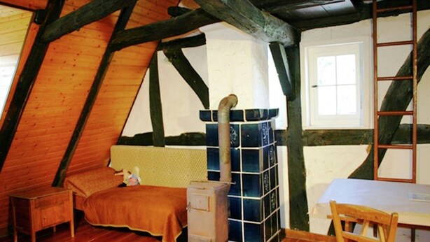 A cozy attic bedroom with wooden beams, a bed, a dresser, and a large window. - Traditionelles Ferienhaus in Hessen mit Garten (Willingshausen)