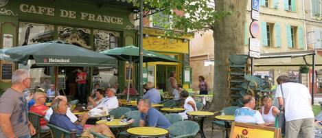 Mobiliario exterior en un café, con mesas y sillas, un letrero que dice Cafe De France y un paraguas de Heineken.