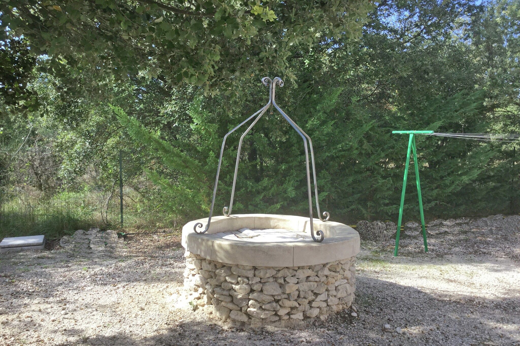A stone well with a metal frame, surrounded by gravel and trees.