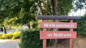 A wooden welcome sign with the text "Willkommen in Nidrum" in a natural setting with trees and a paved path.