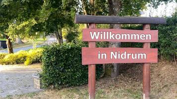 A wooden welcome sign with the text "Willkommen in Nidrum" in a natural setting with trees and a paved path.